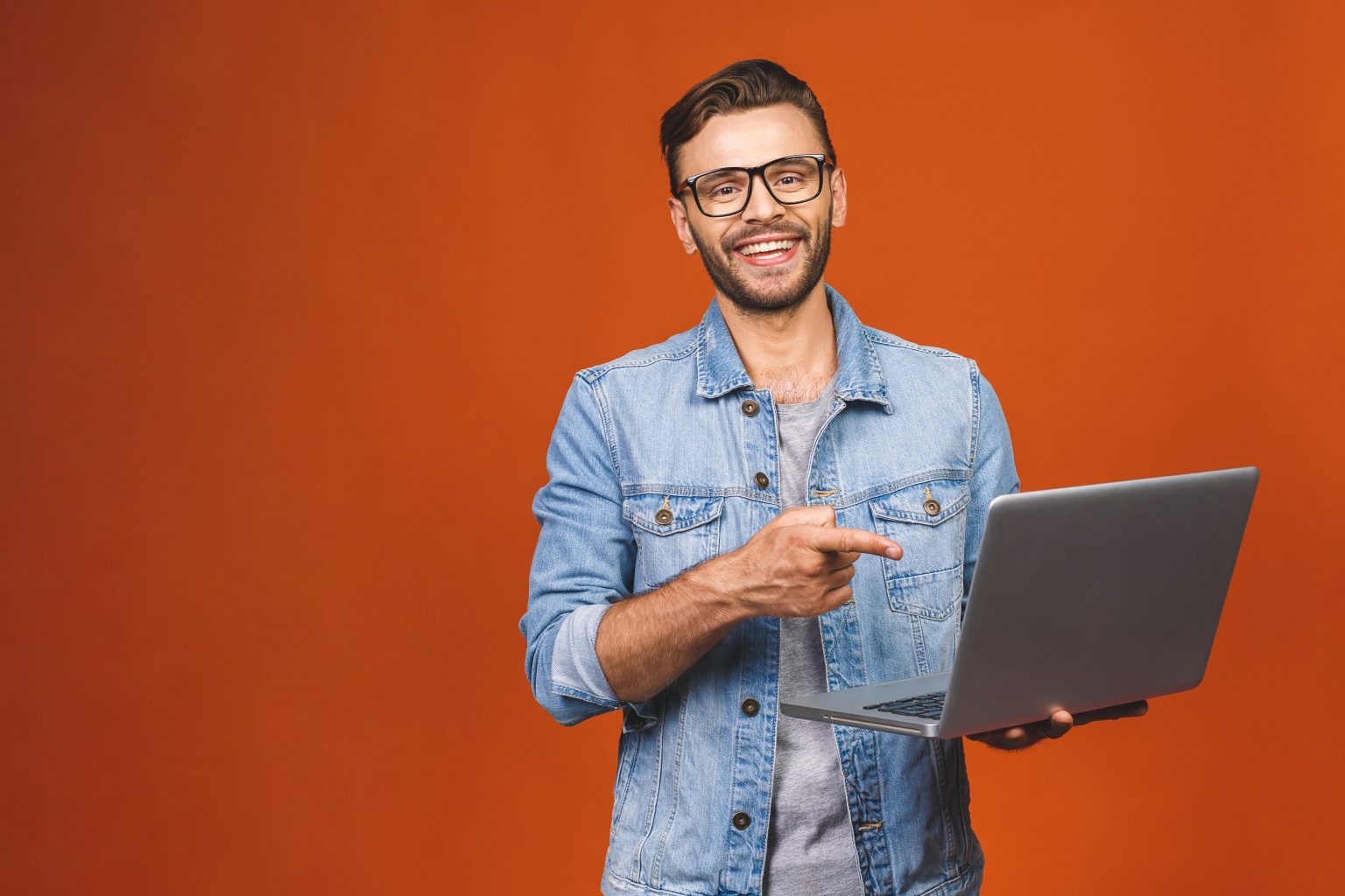 Indoor Portrait Of Young Man In Casual Standing Against Orange Backgroundl With Copy Space For Ads Holding Laptop Smiling