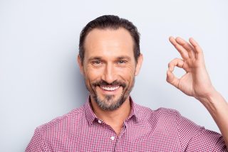 Portrait of good-looking man look at camera stand isolated on light gray background give ok sign make hollywood white smile