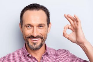 Portrait of good-looking man look at camera stand isolated on light gray background give ok sign make hollywood white smile