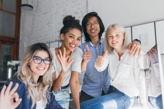 Asian male student spending time with university mates and posing in room with loft interior. Elegant female office worker in white shirt sits on table and having fun with co-workers.