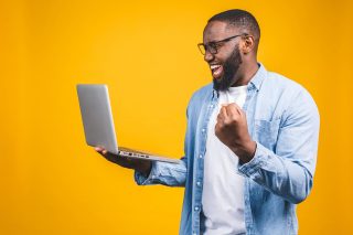 Excited happy afro american man looking at laptop computer screen and celebrating the win isolated over yellow background.