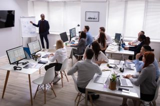 African Businessman Giving Presentation To His Colleagues Sitting On Table With Computers In Office